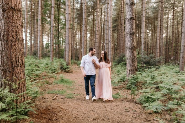 Mum and dad are walking hand in hand and looking at each other. maternity photo shoot in the forest. Mum is wearing pink dress and dad is wearing white shirt and jeans. Maternity photo shoot in Berkshire. Berkshire maternity photographer. Ewa Jones Photography