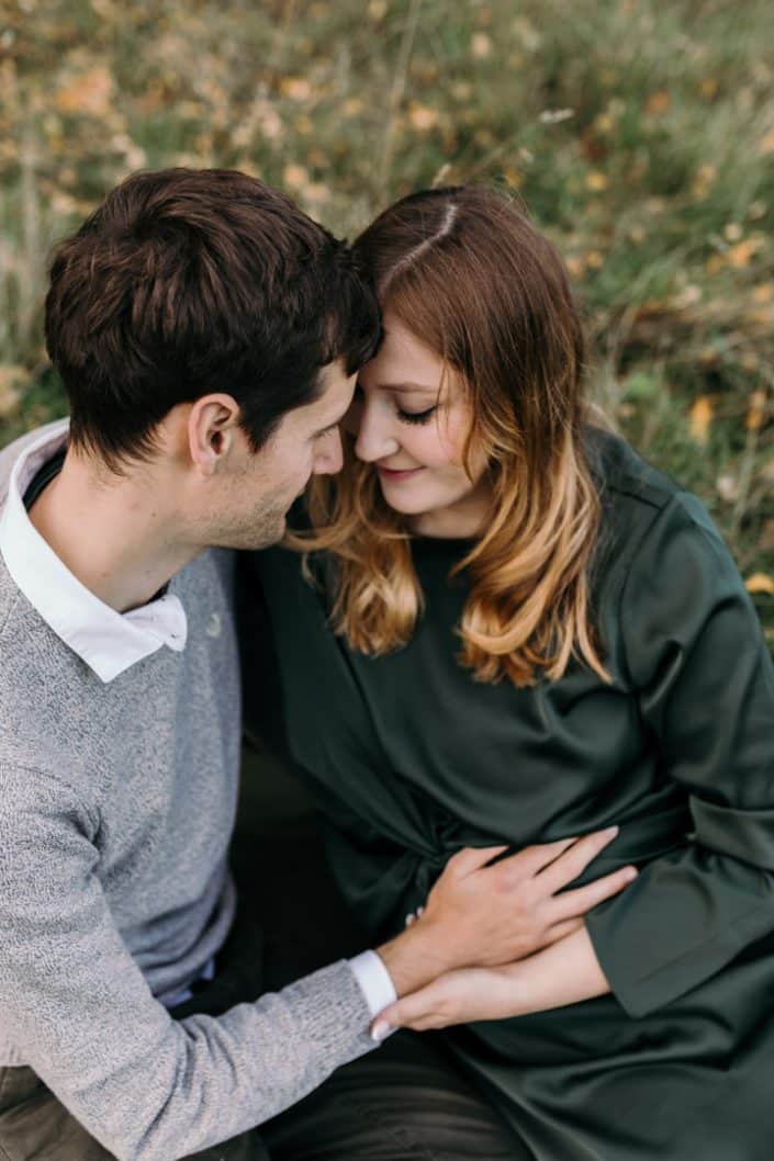 Mum and dad to be are sitting on the grass and looking down. Dad is holding baby bump. Intimate maternity shoot. Maternity pose. Hampshire maternity photo session. Ewa Jones Photography