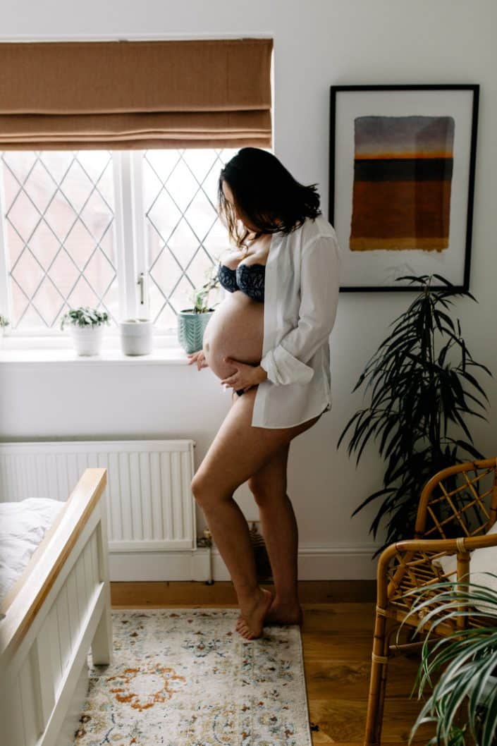 Mum is standing against the window and looking down at her baby bump. she is wearing underwear and white shirt. Hampshire photo shoot at home. Hampshire maternity photographer. Ewa Jones Photography