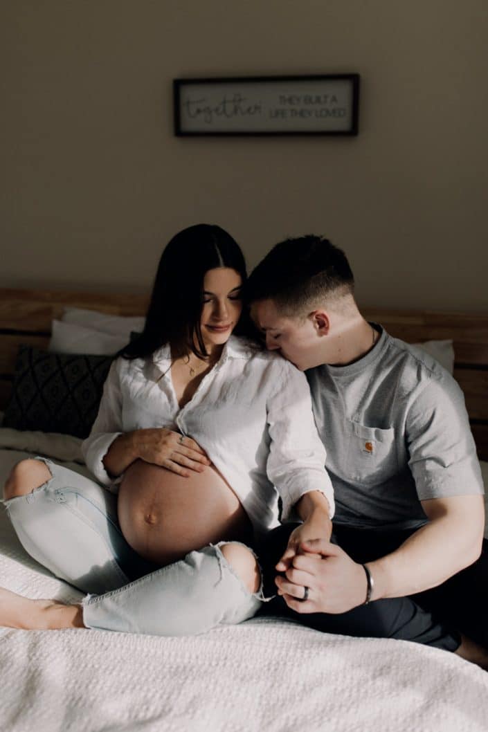 Mum to be and dad are sitting on the bed and dad is kissing mum on her shoulder. Pregnancy photo shoot at home. Hampshire photographer. Ewa Jones Photography