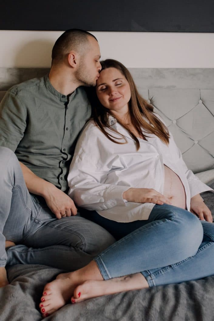 Mum and dad are sitting on the bed and cuddling. Dad is kissing mum in her forehead. Maternity photo shoot at home. Basingstoke maternity photo session. Ewa Jones Photography