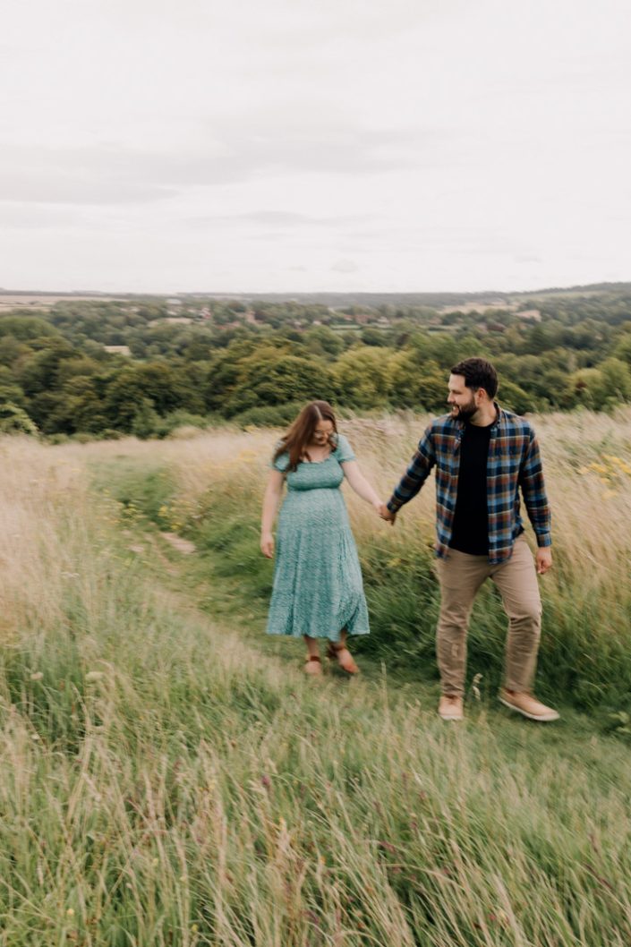 Dad is leading expecting mum. She is looking down and smiling. Maternity photo session in the fields. Hampshire photographer. Ewa Jones Photography