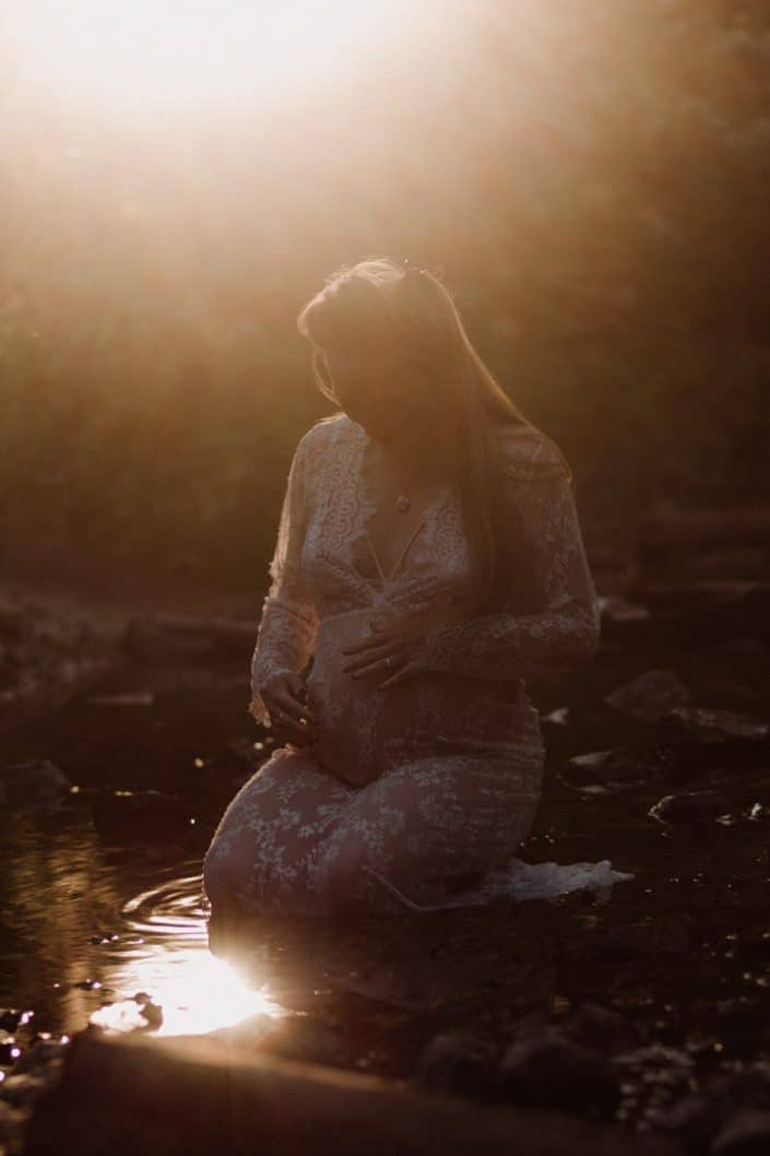 Mother to be is sitting in the river and looking down at her baby. She has a lovely dress. Intimate maternity session. Hampshire photographer. Ewa Jones Photography