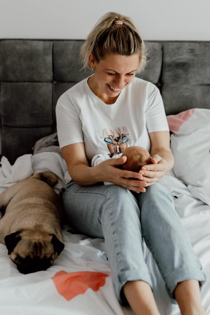 Mum is sitting on the bed and looking down at her baby girl. she is smiling at her girl. Next to mum is laying their dog. Newborn lifestyle photography in Hampshire. Ewa Jones Photography